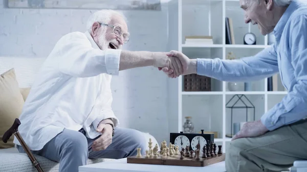 Happy senior men shaking hands near chessboard at home — Stockfoto