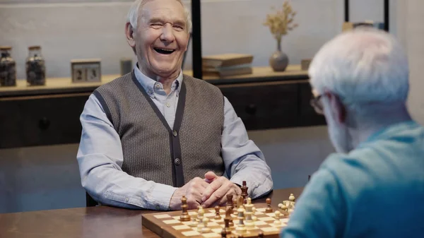 Excited elderly man laughing near chessboard and blurred friend — стоковое фото