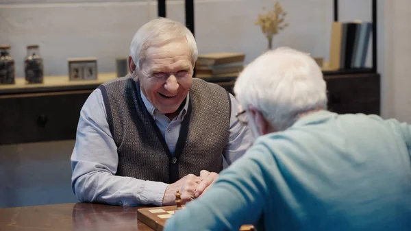 Joyful man looking at senior friend while playing chess at home — Fotografia de Stock