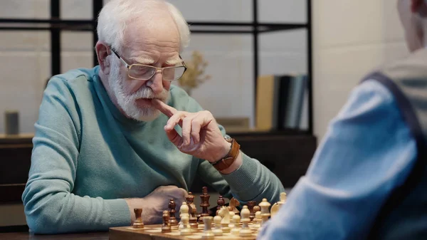 Thoughtful senior man in eyeglasses touching lips while playing chess with blurred friend — Stockfoto