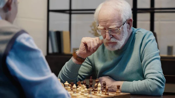 Senior man in eyeglasses thinking near chessboard and blurred friend at home — стоковое фото