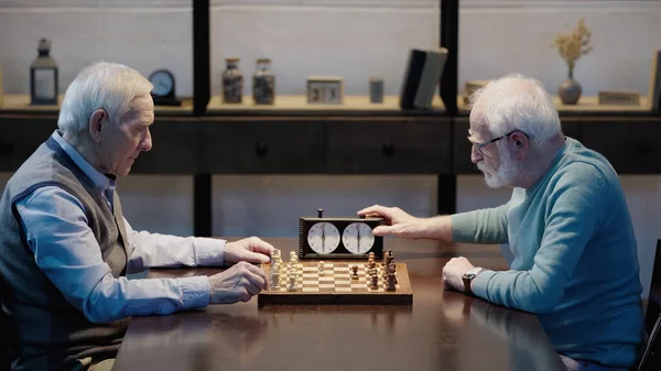 Side view of bearded senior man fixing time on chess clock while playing with friend at home — Photo de stock