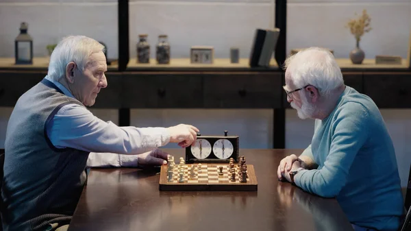 Side view of senior man fixing time on chess clock near friend in living room - foto de stock