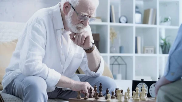 Senior grey haired man in eyeglasses playing chess with friend in living room — Fotografia de Stock