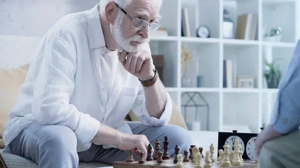 Senior bearded man thinking near chessboard and blurred friend - foto de stock