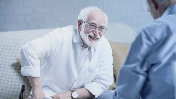 Cheerful senior man in eyeglasses laughing near blurred friend at home — Stock Photo