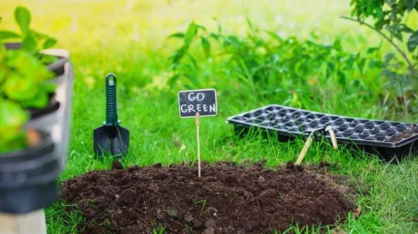 Board with go green lettering in soil near gardening tools and grass in garden — Stock Photo