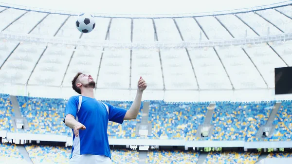 Football player in blue t-shirt bouncing ball with head on stadium — Stock Photo