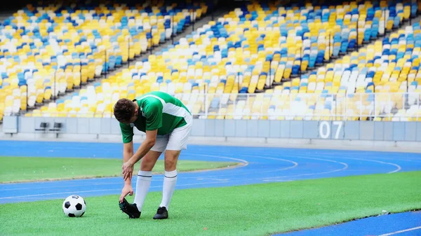 Football player in green uniform stretching near ball of field in stadium — стоковое фото