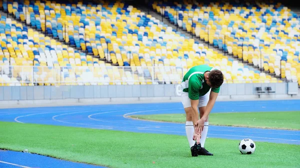 Young football player in uniform stretching near ball of green field — стоковое фото