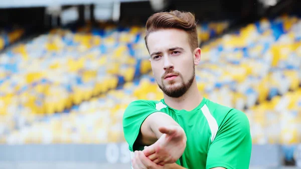 Bearded football player in t-shirt stretching hands while warming up on stadium — Fotografia de Stock