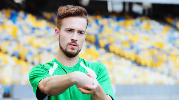 Young football player in t-shirt stretching hands while warming up on stadium — Stockfoto