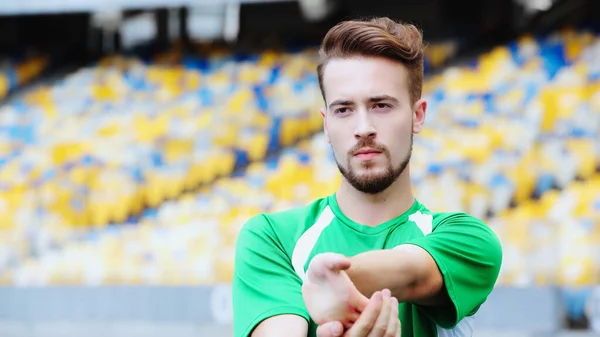 Football player in green t-shirt stretching hands while warming up on stadium - foto de stock