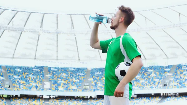 Side view of young football player in green t-shirt holding ball while drinking water — Stockfoto