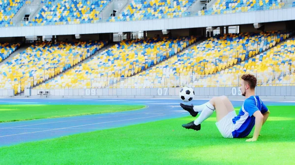 Side view of football player sitting on green grass and bouncing ball with legs — Fotografia de Stock