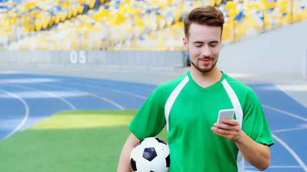 Smiling football player holding ball and messaging on smartphone — стоковое фото