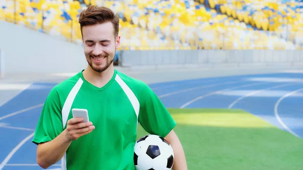 Smiling football player holding ball and messaging on smartphone — Stockfoto
