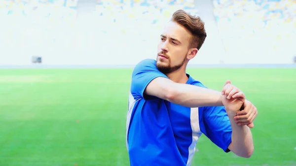 Young football player in blue t-shirt warming up on green field of stadium — Foto stock