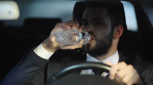 Thirsty businessman driving car and drinking pure water from plastic bottle — Stock Photo