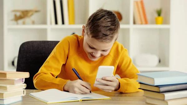 Happy schoolboy using smartphone while writing in notebook near books on desk — стоковое фото