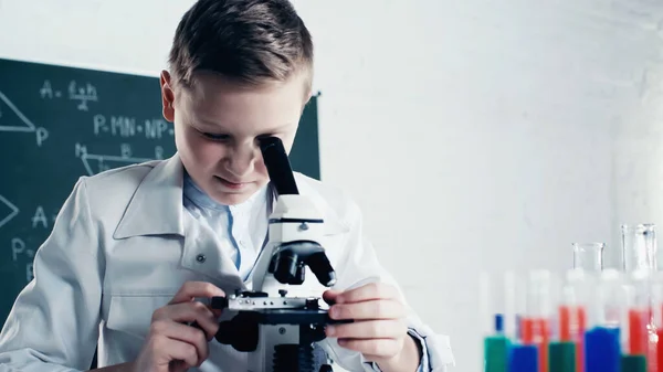 Школярка в білому пальто дивиться через мікроскоп під час уроку в класі — стокове фото