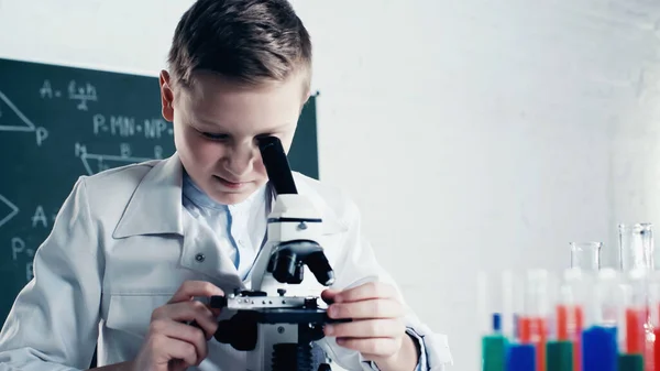 Schoolkid in white coat looking through microscope during lesson in classroom — Stock Photo