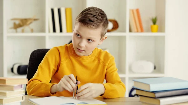 Schoolboy in yellow jumper using drawing compass near books on desk — Fotografia de Stock