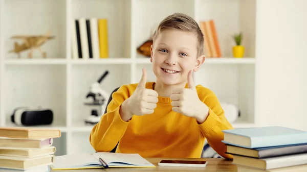 Happy schoolboy showing thumbs up near smartphone and books on desk — Stock Photo