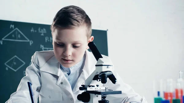 Schoolboy in white coat writing near microscope during chemistry lesson in classroom — Stock Photo