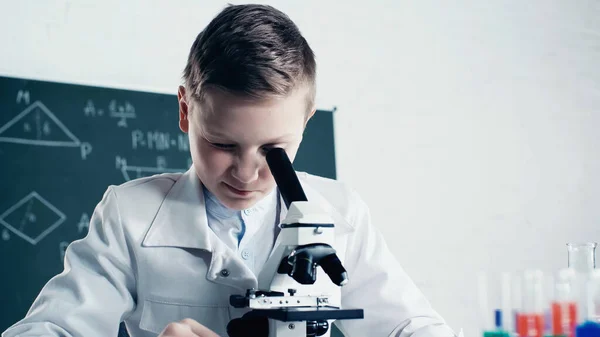 Хлопчик у білому пальто дивиться через мікроскоп під час уроку хімії в класі — стокове фото