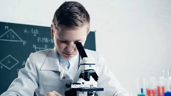 Boy in white coat looking through microscope during chemistry lesson in classroom — Stock Photo