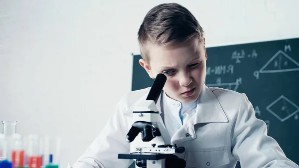 Schoolboy in white coat looking through microscope during chemistry lesson in classroom — Stock Photo