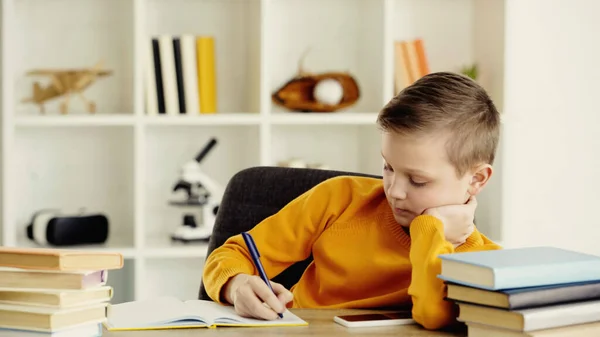 Schoolboy writing in copy book near smartphone with blank screen and books on desk — Stock Photo