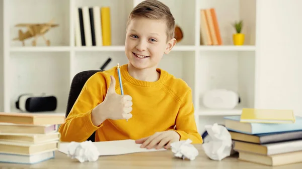 Cheerful schoolboy smiling and showing thumb up near books and crumpled papers at home — Stock Photo