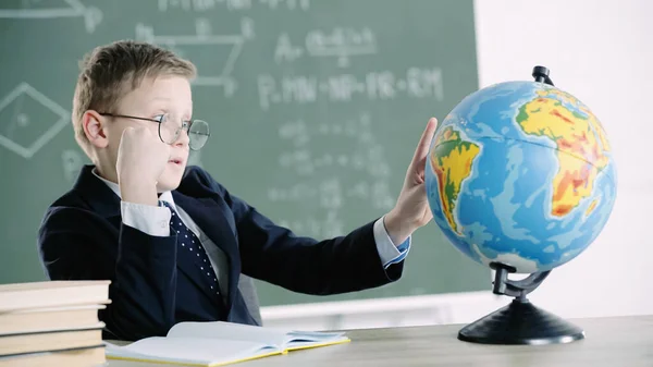 Schoolboy in glasses pointing with finger at globe while sitting near books in classroom — Stock Photo