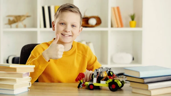 Happy schoolboy sitting near toy car made of building blocks and showing thumb up at home — стоковое фото