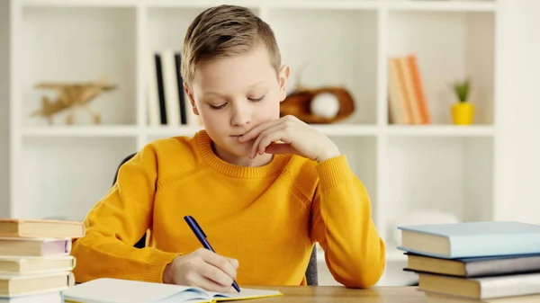 Thoughtful preteen boy writing in notebook while doing homework near books on wooden desk — стоковое фото