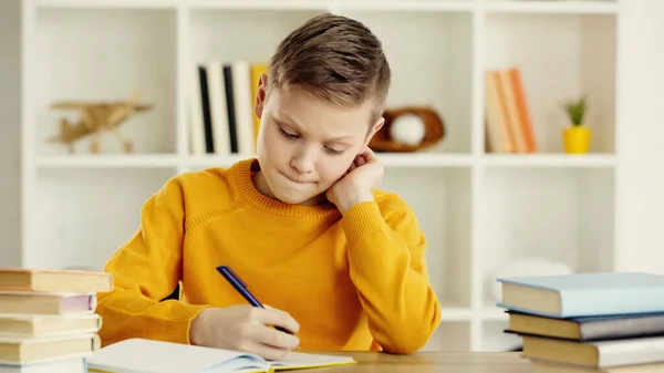 Thoughtful preteen schoolkid writing in notebook while doing homework near books on wooden desk — Stock Photo