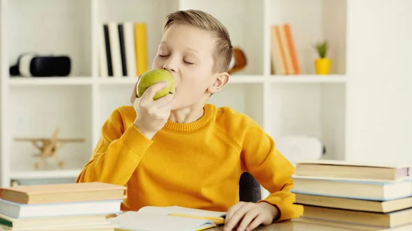 Schoolboy with closed eyes biting green and tasty apple near books at home — Stock Photo