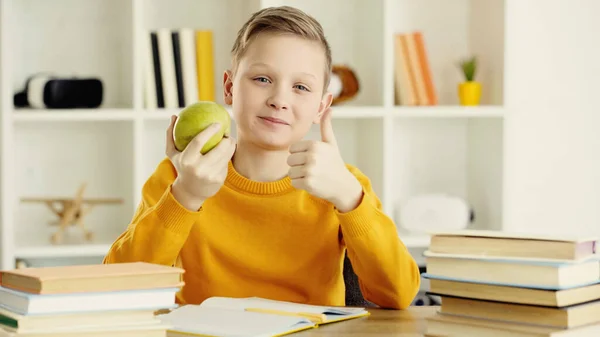Schoolboy holding green and tasty apple while showing thumb up at home — Stock Photo