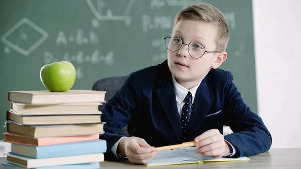 Schoolboy in glasses holding pen near notebook and looking at apple on books in classroom — Stock Photo