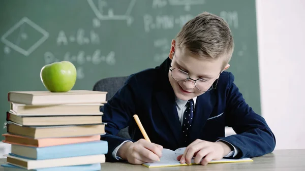 Cheerful schoolboy in glasses writing in notebook near apple on books in classroom — Stock Photo