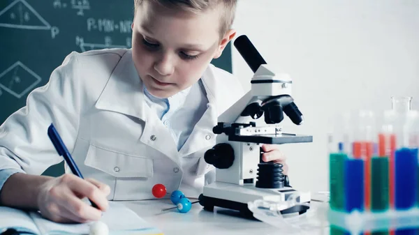 Schoolkid in white coat making notes near microscope during lesson in classroom — Stock Photo