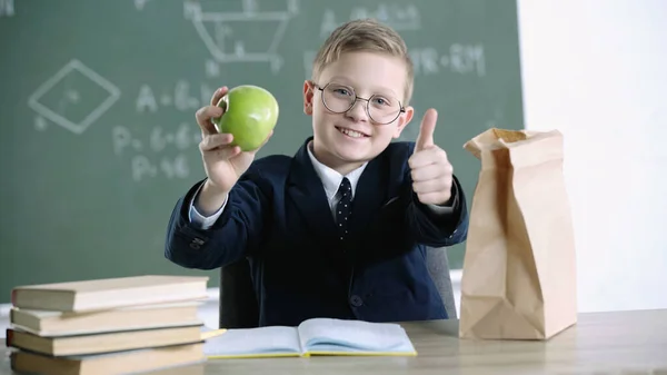 Happy schoolboy in glasses holding green apple while showing thumb up near paper bag with lunch in school — Stock Photo
