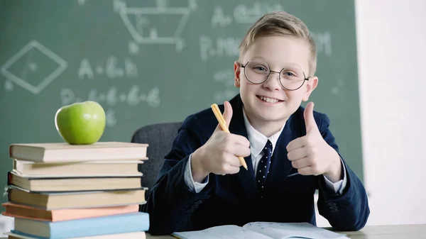 Pleased schoolboy in glasses showing thumbs up near apple and books in school — Stock Photo