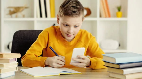 Schoolboy looking at smartphone while writing in notebook near books in classroom — Stock Photo