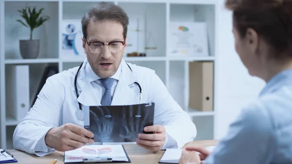 Serious doctor sitting at workplace while looking at x-ray scan near woman on blurred foreground — Fotografia de Stock