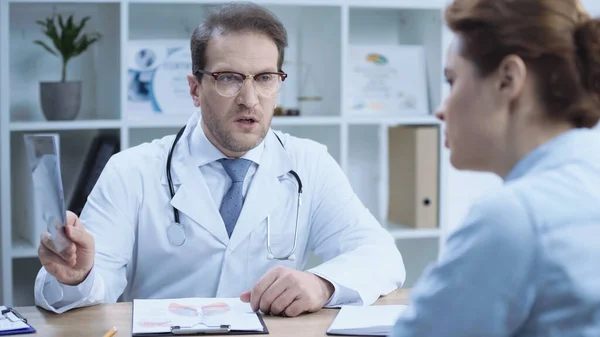 Serious doctor holding x-ray scan while talking with patient on blurred foreground — стоковое фото