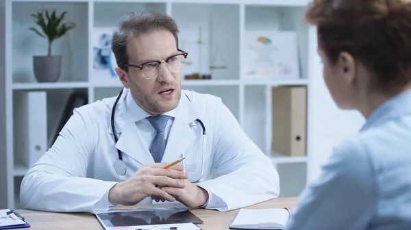 Serious doctor in glasses and white coat sitting with clenched hands while talking with patient on blurred foreground - foto de stock