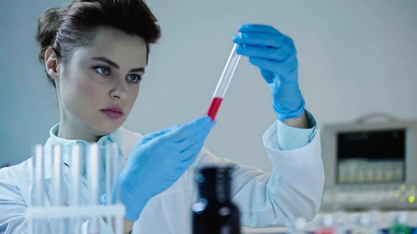 Attentive scientist looking at red sample in test tube near blurred medical equipment — Photo de stock